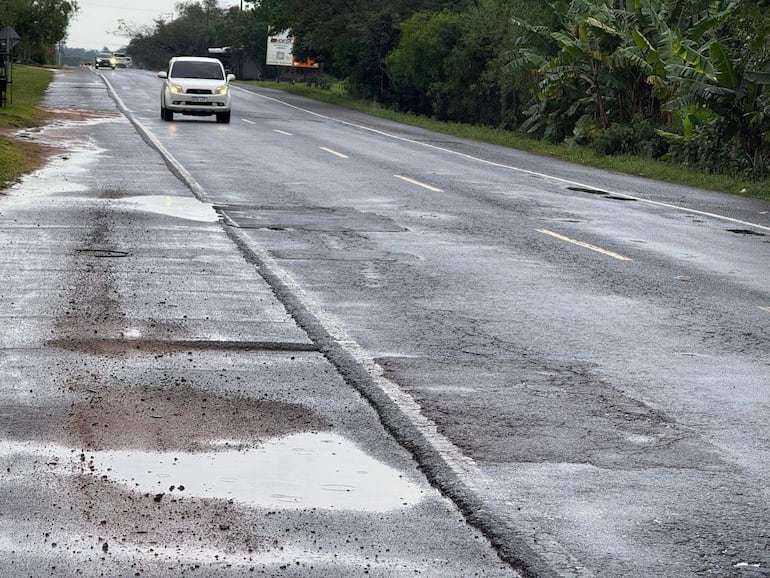 Carretera deteriorada rodeada de vegetación, con un automóvil blanco avanzando y otro vehículo a lo lejos.