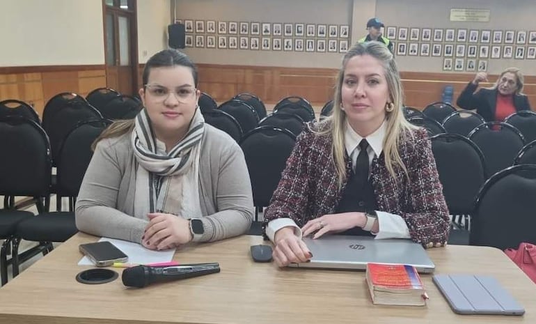 Belinda Bobadilla con bufanda y suéter y Mara Gómez con abrigo de cuadros se preparan para hablar en un salón vacío.