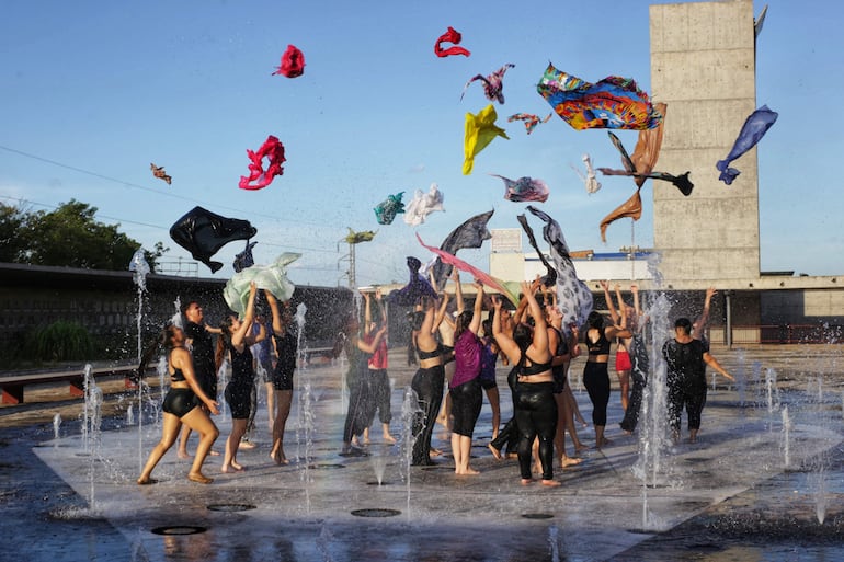 Veinte personas, en ropa deportiva oscura, coreografían lanzamiento de telas de colores en la plaza, rodeadas de chorros de agua.