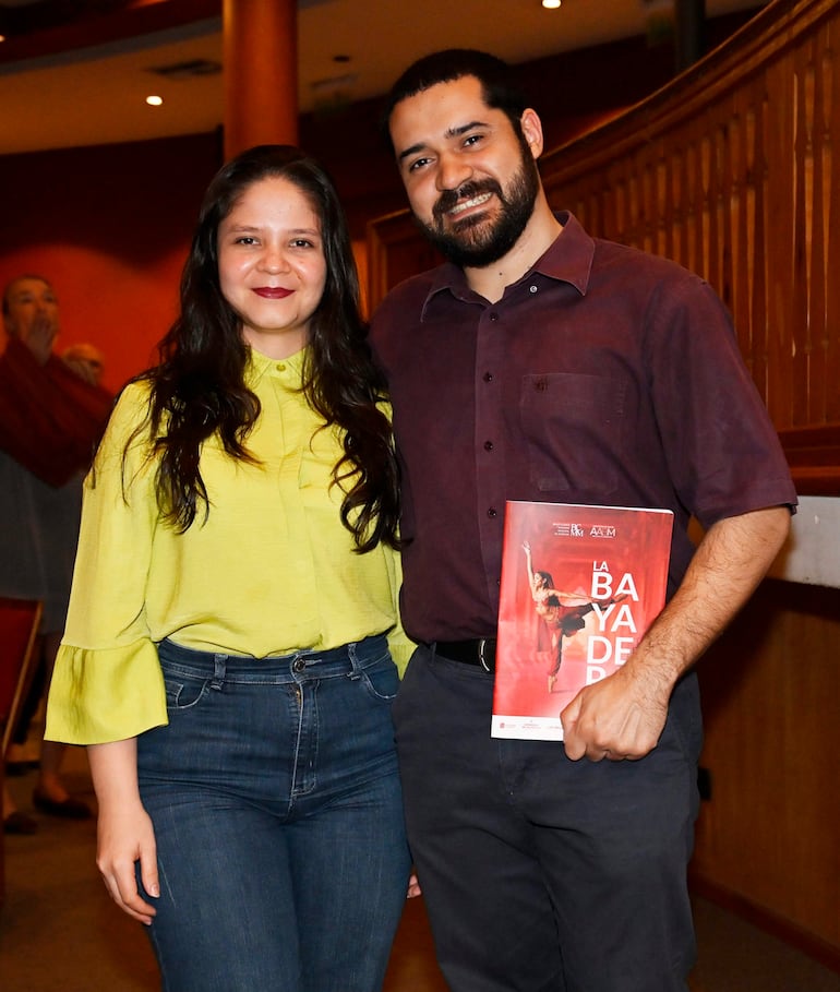 Mujer con blusa amarilla y jeans, y hombre con camisa marrón oscuro sosteniendo un folleto, sonríen en un ambiente acogedor.