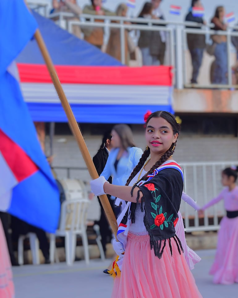 Niña con falda rosa y blusa blanca sostiene un estandarte en un desfile, rodeada de personas con banderas.
