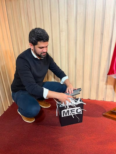 Raúl Benítez en suéter negro y jeans, inclinado frente a una caja negra con inscripción 'MEC', en un ambiente formal.