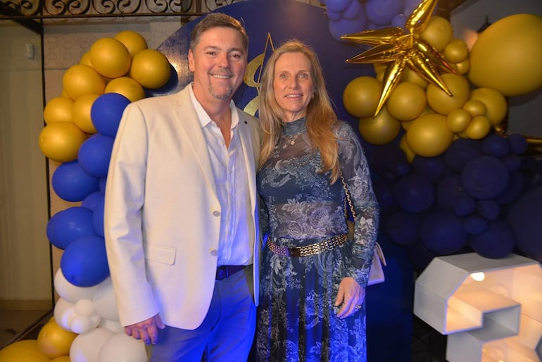 Pareja sonriente: hombre con chaqueta clara y mujer en vestido oscuro con diseño floral, rodeados de globos decorativos.