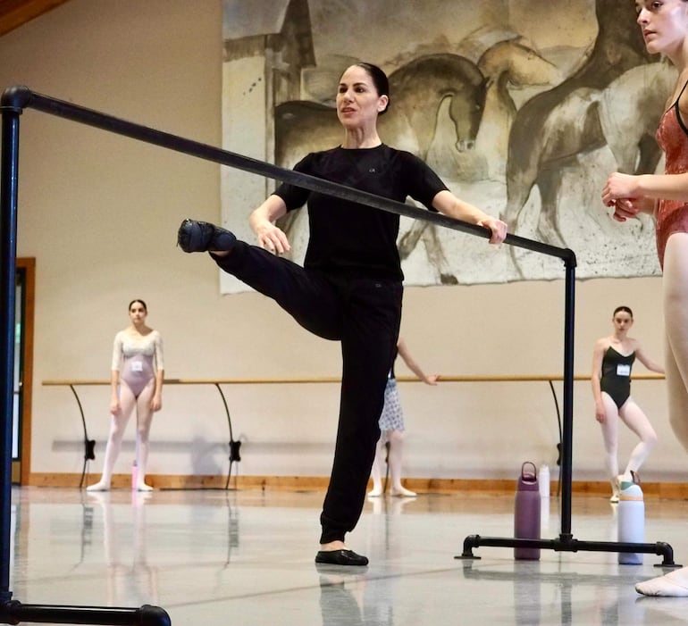 Mujer con cabello recogido y vestimenta negra muestra técnica de ballet, rodeada de bailarinas en diversas posturas.