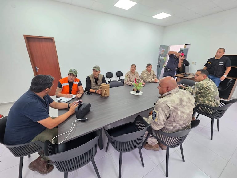 Hombre con camiseta negra sostiene un teléfono y un bolso, mientras joven con chaqueta naranja toma notas en una reunión interior.
