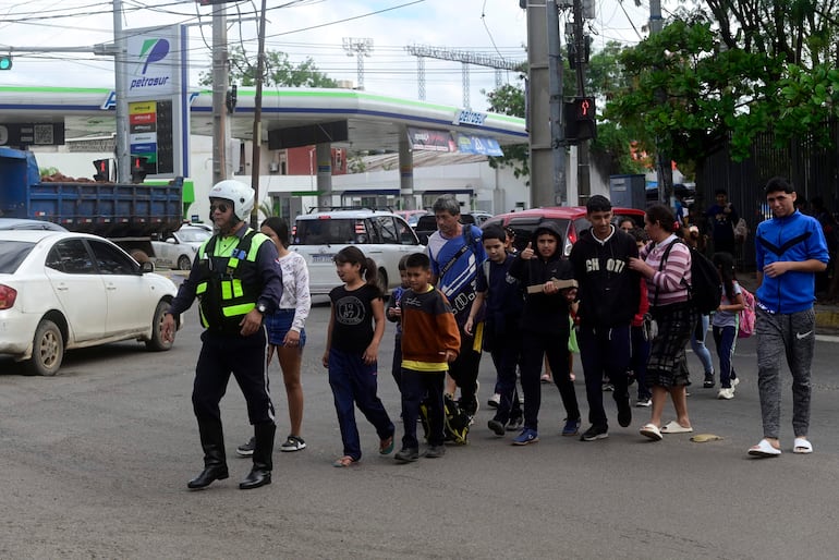 Oficial de policía con chaleco reflectante guía a jóvenes sonrientes cruzando la calle, con vehículos al fondo.