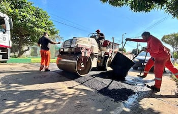 Tres hombres trabajando en el asfalto: uno con camiseta oscura supervisa, otro con conjunto rojo sostiene un palín, y un tercero opera una maquinaria.