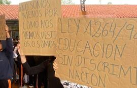 Grupo de estudiantes con carteles en un aula, uno en camiseta oscura levanta el brazo, otro sostiene un gran cartel sobre educación.