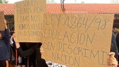 Grupo de estudiantes con carteles en un aula, uno en camiseta oscura levanta el brazo, otro sostiene un gran cartel sobre educación.