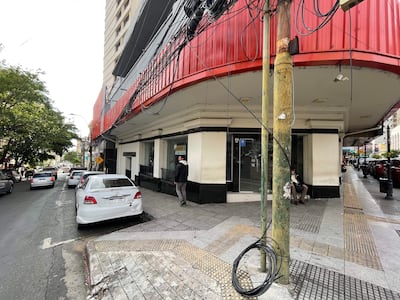 Hombre en chaqueta negra camina junto a otro sentado en silla, en un ambiente urbano vacío y melancólico.