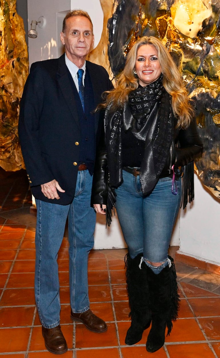 Hombre canoso en blazer oscuro y mujer rubia en abrigo negro, sonríen juntos en interior con arte abstracto de fondo.