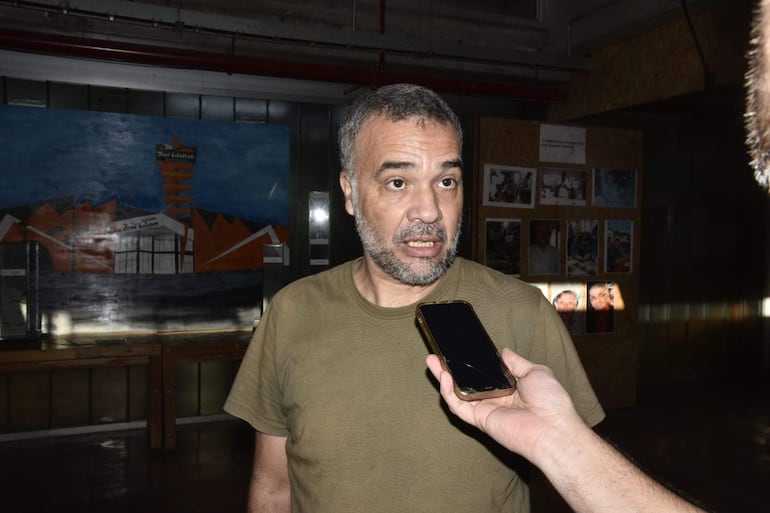 Hombre de barba corta y cabello gris con camiseta oliva habla, mientras otra persona sostiene un teléfono móvil en un ambiente iluminado.