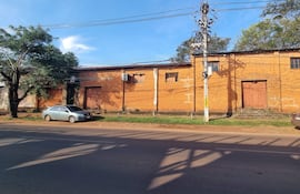 Terreno con dos vehículos estacionados y un árbol frondoso, frente a una pared de ladrillos naranjas.