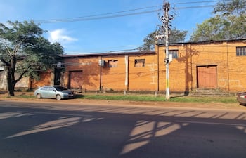 Terreno con dos vehículos estacionados y un árbol frondoso, frente a una pared de ladrillos naranjas.