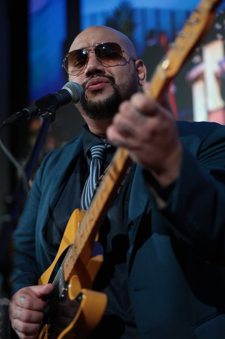 Hombre con traje azul y gafas de sol sostiene guitarra eléctrica amarilla en un escenario.