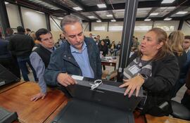 Hombre canoso con chaqueta oscura observa dispositivo, mientras una mujer con chaqueta negra gesticula explicando en reunión interna.