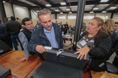 Hombre canoso con chaqueta oscura observa dispositivo, mientras una mujer con chaqueta negra gesticula explicando en reunión interna.