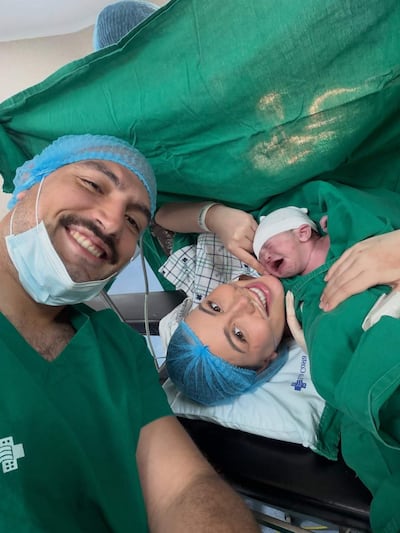 Padre sonriente en camiseta verde quirúrgica sostiene a su recién nacido junto a la madre en bata y gorro quirúrgico.