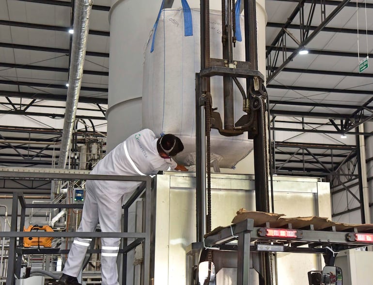 Hombre en camisa blanca y pantalones claros ajusta un recipiente en un entorno industrial con estructuras metálicas visibles.