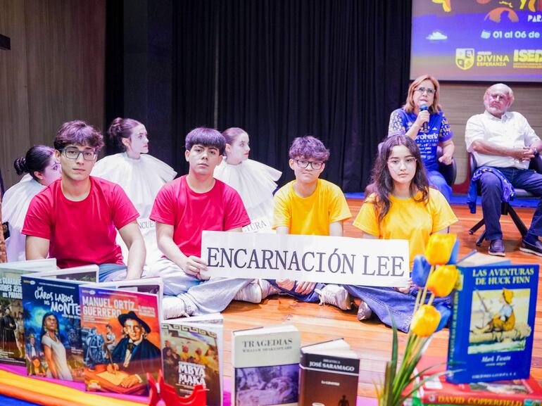 Cuatro jóvenes sentados en el suelo con camisetas rojas y una amarilla sosteniendo un cartel, rodeados de libros y un ambiente colorido.