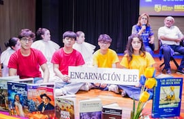 Cuatro jóvenes sentados en el suelo con camisetas rojas y una amarilla sosteniendo un cartel, rodeados de libros y un ambiente colorido.