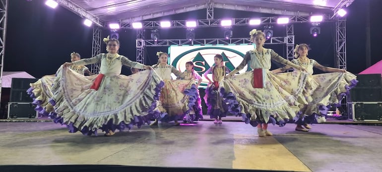 Seis bailarinas con trajes tradicionales de colores claros y púrpuras realizan una coreografía en un escenario iluminado.