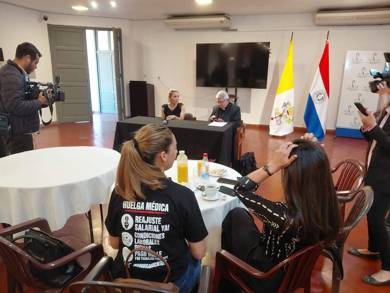 Adalberto Martínez con gafas escucha atentamente a Rossana González, quien habla frente a un micrófono en un ambiente istitucional.