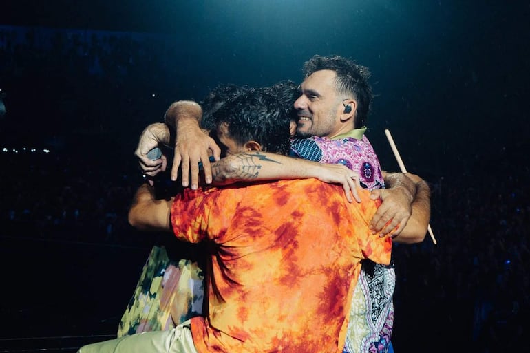 Tres personas abrazándose en el escenario, una con micrófono y otra con camiseta naranja, todos sonrientes y felices.