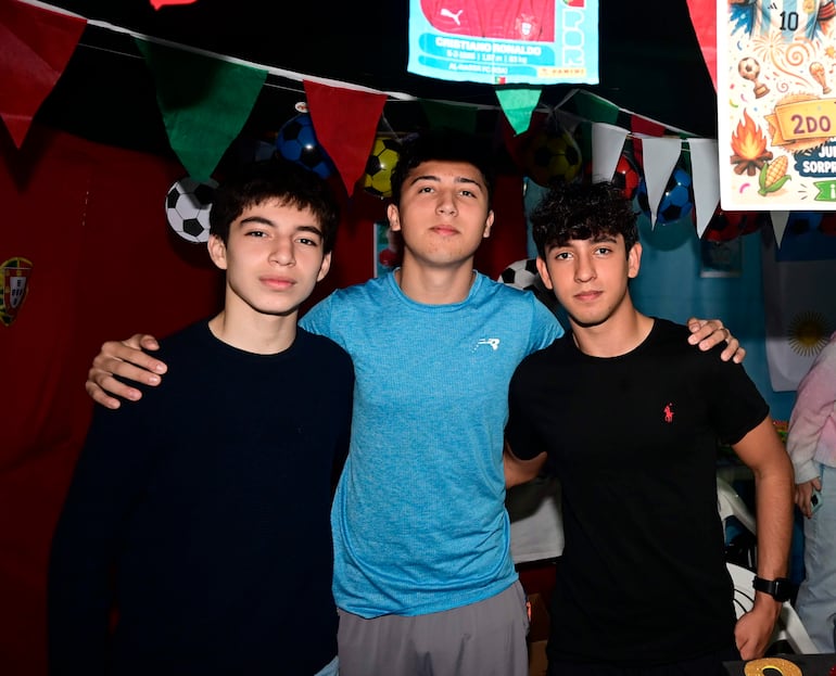 Tres jóvenes sonrientes, uno en camiseta negra y dos en azul y negro, rodeados de decoraciones futbolísticas.