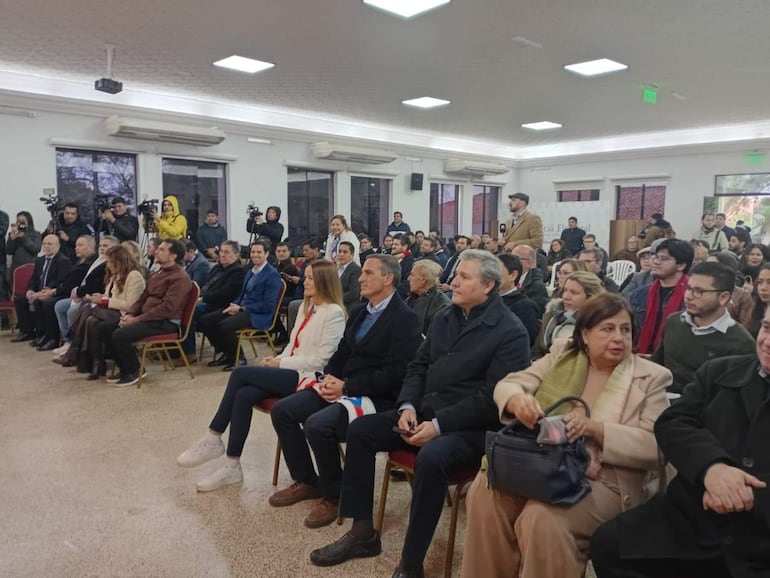 Soledad Núñez con abrigo blanco, rodeada de personas en un evento formal con actitud de escucha y camarógrafos al fondo.