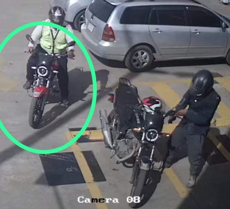 Hombre con chaleco reflectante en motocicleta roja sentado mientras otro hombre con casco ajusta una motocicleta negra en un estacionamiento.