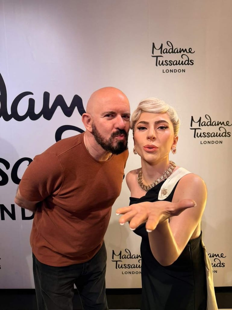 Hombre calvo con barba en camiseta marrón e inclinación hacia una mujer rubia en vestido negro en Madame Tussauds.