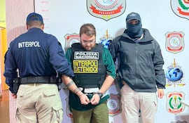Hombre esposado en el centro, vestido con chaleco 'INTERPOL DETENIDO', flanqueado por dos hombres en un ambiente policial tenso.