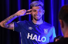 Julio Enciso sonriente, vistiendo camiseta azul de Ipswich Town, hace signo de paz con la mano en un fondo oscuro.
