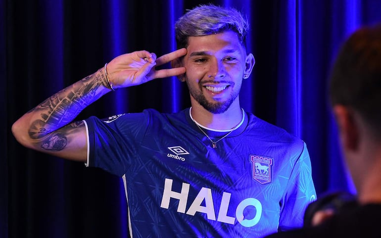 Julio Enciso sonriente, vistiendo camiseta azul de Ipswich Town, hace signo de paz con la mano en un fondo oscuro.