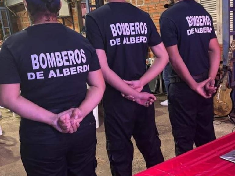 Tres personas de espaldas con camisetas negras que dicen 'BOMBEROS DE ALBERDI', en un interior con paredes de ladrillo.