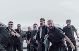 Siete hombres con camisetas oscuras y pantalones cortos, sonriendo y moviéndose con alegría en un ambiente festivo exterior.