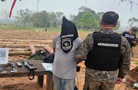 Detenido con capucha negra es custodiado por agente en chaleco, rodeados de evidencias en terreno rural desforestado.