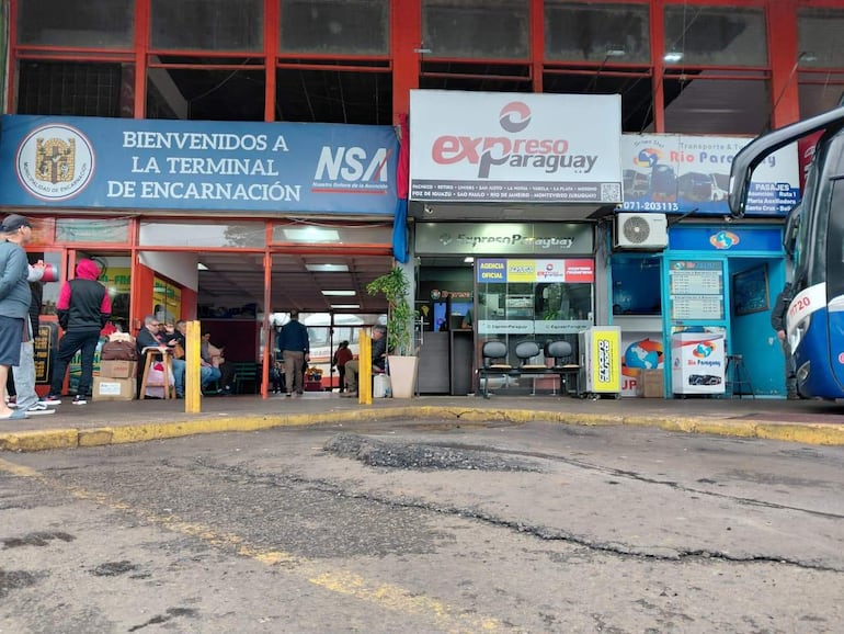 Hombres sentados en mesas en la Terminal de Encarnación, unos en pantalones cortos, otros con abrigo oscuro, en un ambiente de espera.