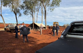 Grupo de seis personas, incluyendo dos policías, en entorno rural con tierra roja, mientras un hombre de gorra blanca habla.