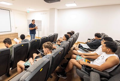 Hombre con gafas y camisa oscura presenta en sala moderna, mientras jóvenes atentos en asientos sostienen tabletas y documentos.