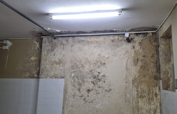 Espacio interior del hospital con paredes deterioradas, manchas de humedad y moho, iluminado por un tubo fluorescente.