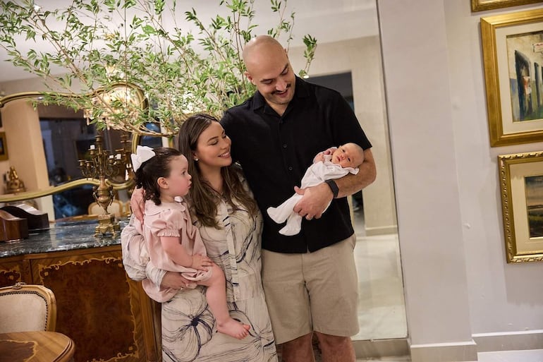 Mujer sonriente con vestido claro sostiene a niña con vestido rosa; hombre feliz abraza a un bebé en un interior elegante.