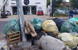 Mujer con ropa azul camina en una calle comercial, rodeada de bolsas de basura y un coche rojo estacionado.