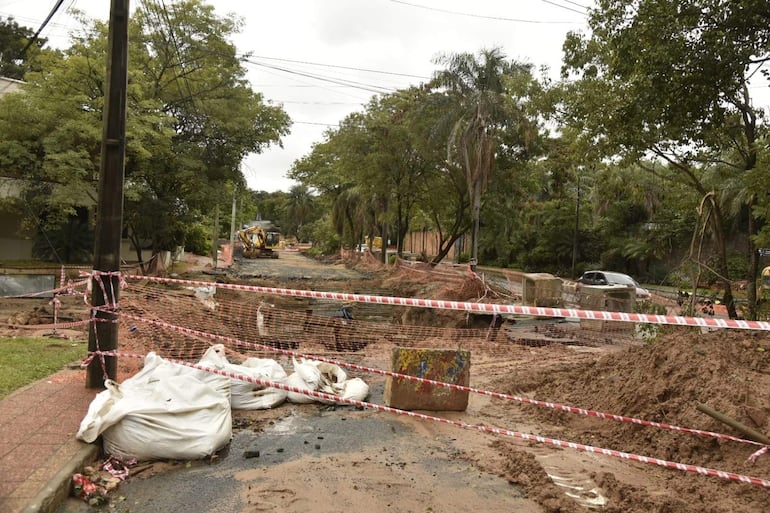 Ambiente de construcción con excavadora amarilla, sacos de arena y cintas de advertencia en un terreno de tierra y lodo.