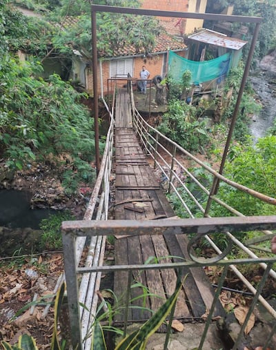Hombre de pie en un puente de madera deteriorado, con vegetación al fondo y edificios visibles detrás.