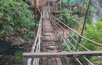 Hombre de pie en un puente de madera deteriorado, con vegetación al fondo y edificios visibles detrás.