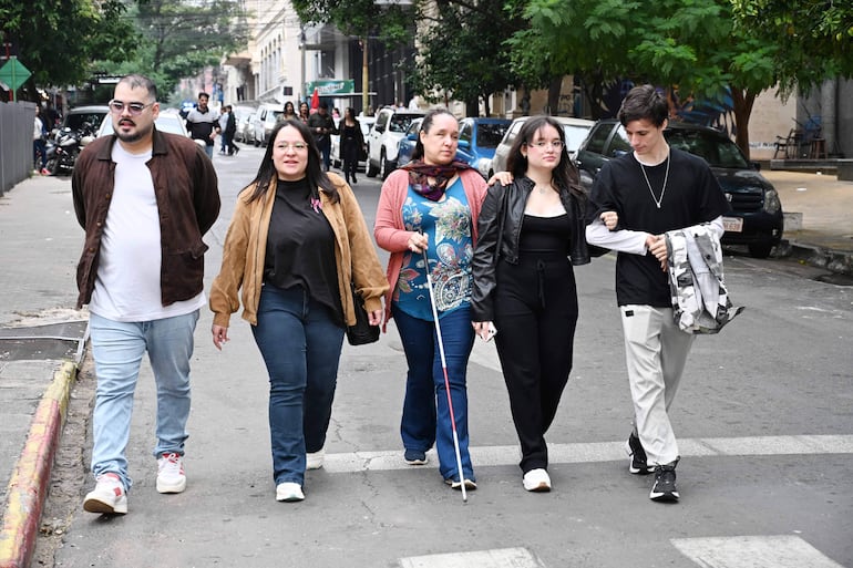 Cinco jóvenes, una chaqueta marrón y un top negro, caminan juntos, conversando en una calle nublada con árboles y vehículos al fondo.