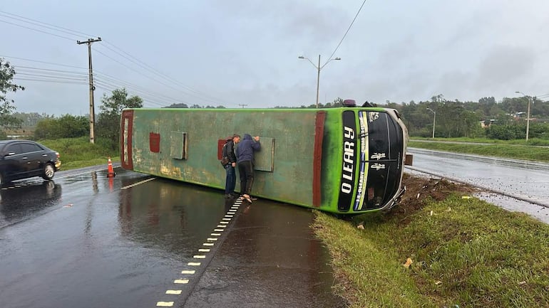 Dos hombres observan un autobús volcado en una carretera mojada; el autobús es verde con la inscripción 'O'Leary'.