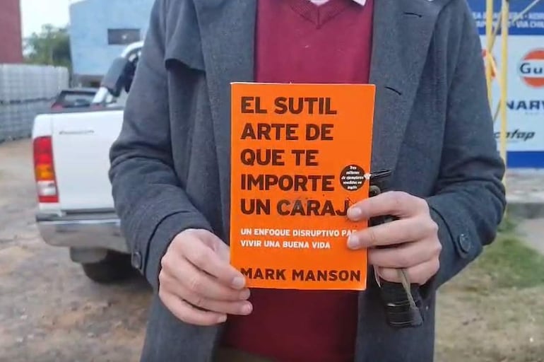 Hombre con chaqueta oscura y suéter rojo sostiene un libro, en un entorno de allanamiento con un vehículo blanco al fondo.